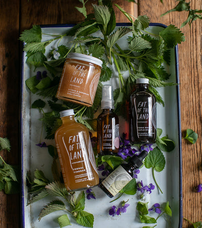 An array of seasonal products, ranging from a body scrub, bitters formula, vinegar, syrup, and mist - are presented atop a tray along with herbs. 