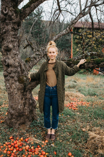 woman stands at a bare apple tree, arms stretched behind her to rest of a bough. apples at her feet. 