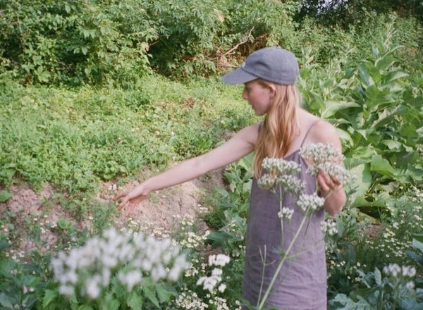a woman, standing in a lush garden, turning away and gesturing toward a plant